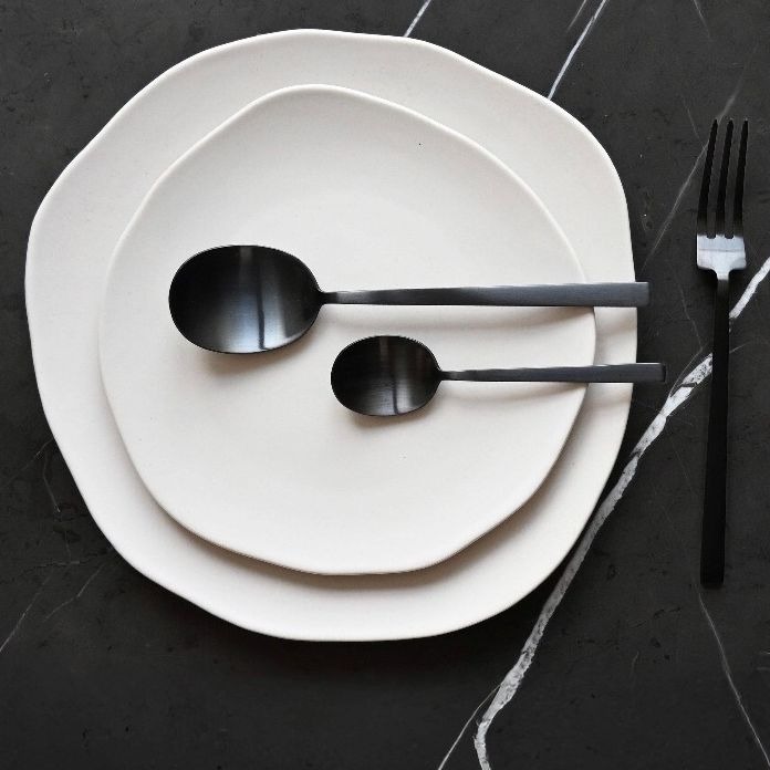 A top-down table setting: two off-white plates holding black spoons, with a black fork and knife laid beside them on a dark surface